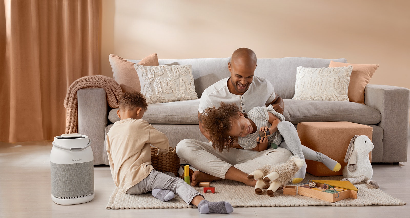 Black father and two multi-racial children playing in front of a couch. Air purifier on floor.