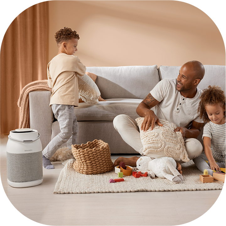 Black father and two multi-racial children playing in front of a couch. Air purifier on floor.